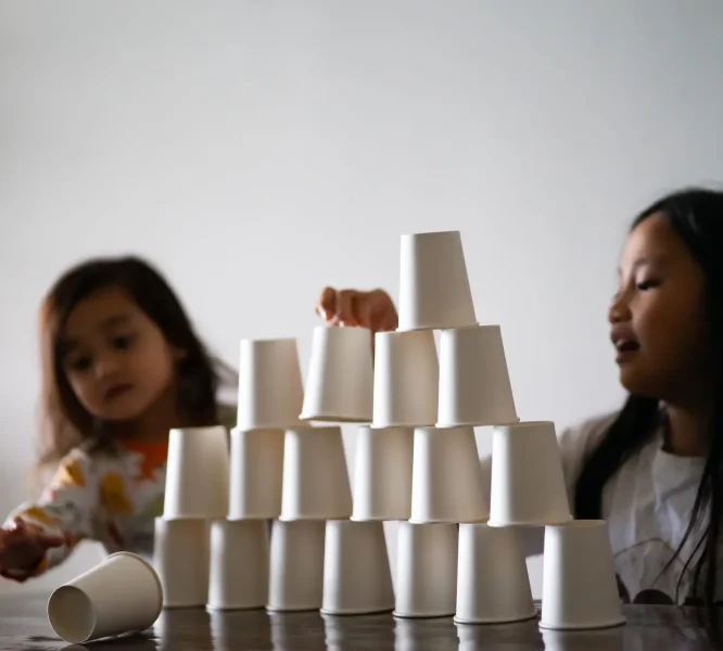 Kids Playing with white paper cups building a cup tower on the table.  Sisters, siblings playing together.